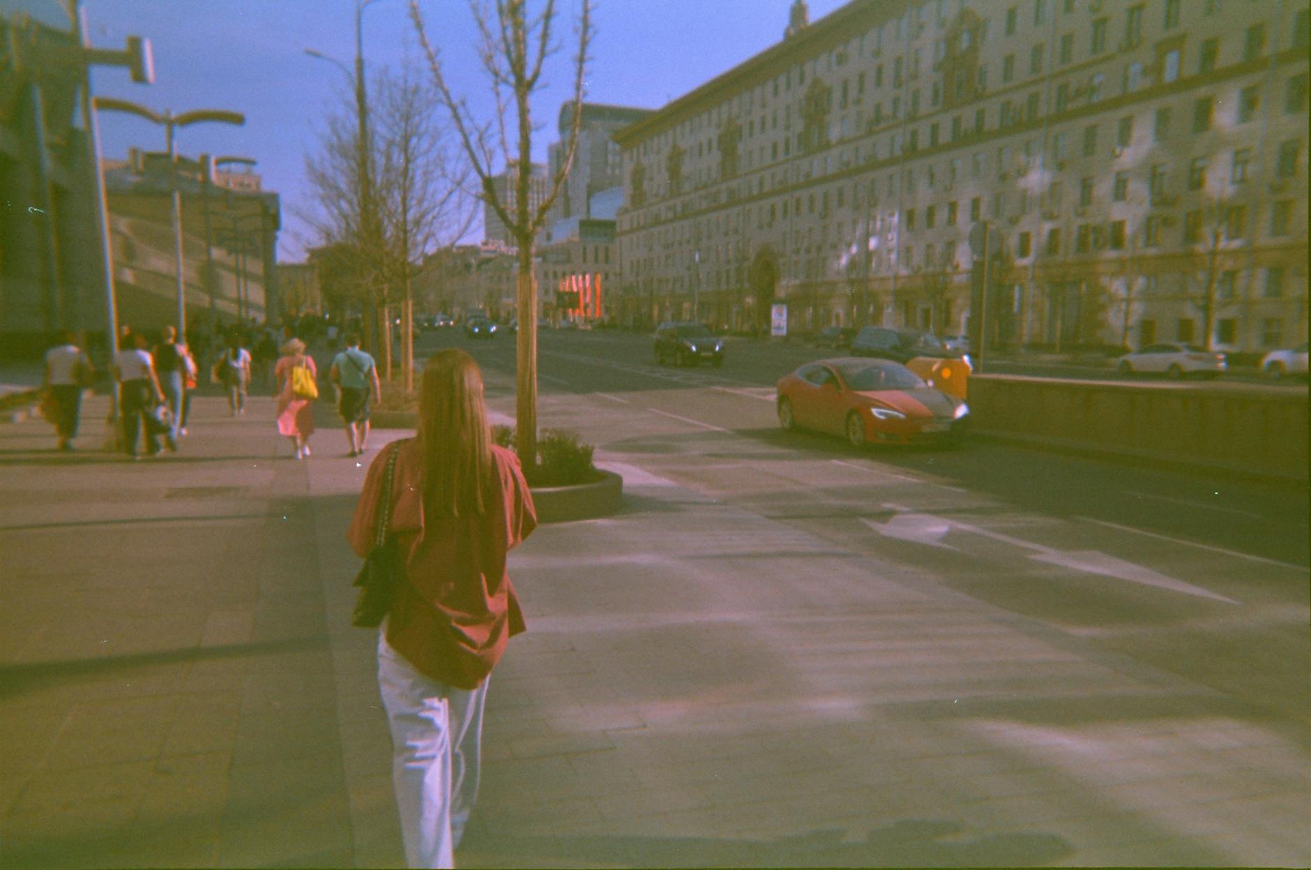 A woman walks along the bustling streets of Moscow with a red car passing by.